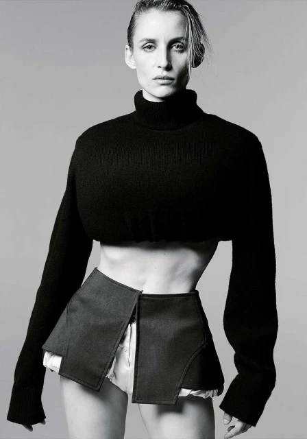Monochrome fashion portrait of a slim adult woman in a cropped black turtleneck sweater and open black skirt, posing in a studio.