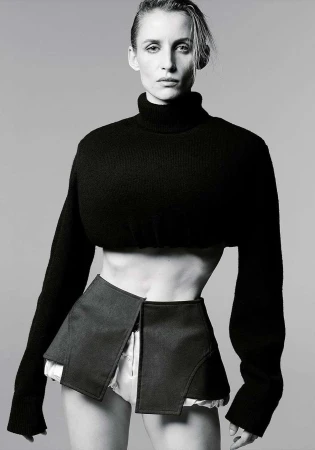 Monochrome fashion portrait of a slim adult woman in a cropped black turtleneck sweater and open black skirt, posing in a studio.