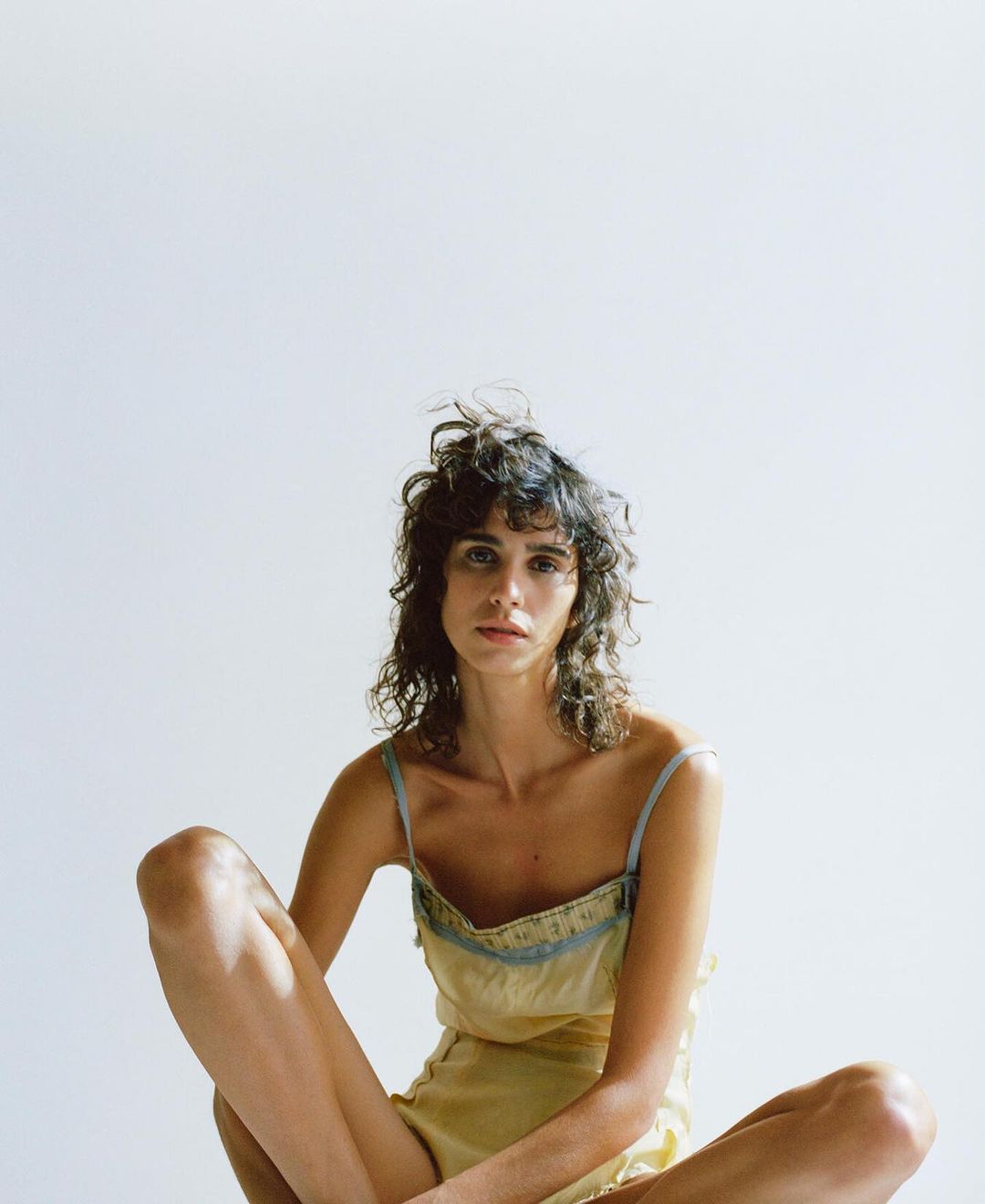 Woman with wavy brown hair sits cross-legged in a yellow camisole and shorts in a studio with white background and soft, diffused lighting.