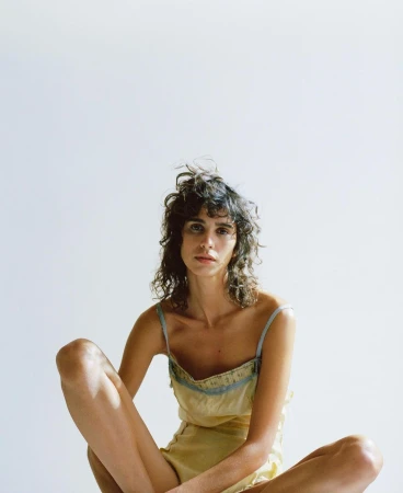 Woman with wavy brown hair sits cross-legged in a yellow camisole and shorts in a studio with white background and soft, diffused lighting.