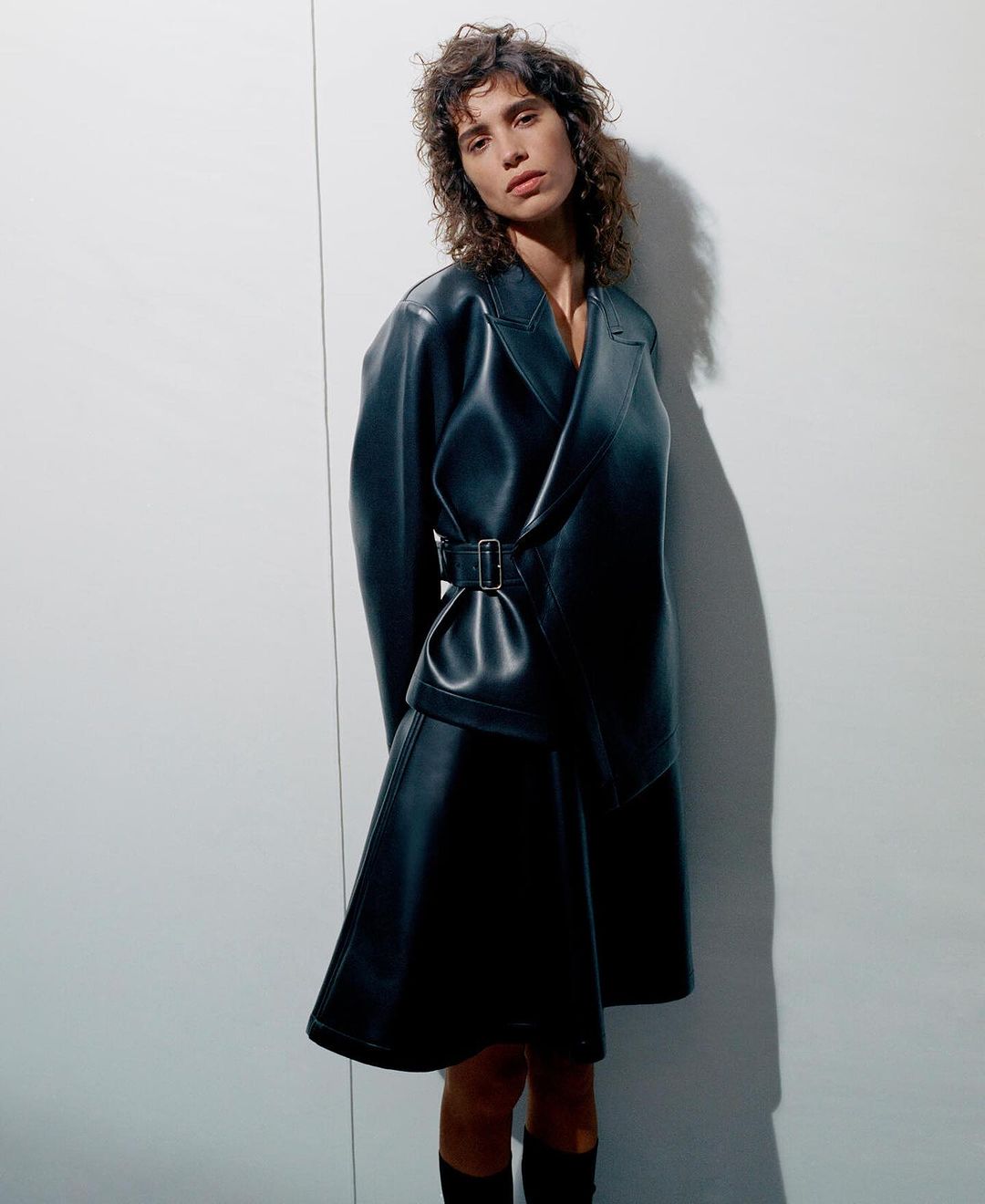 A woman in a black leather belted dress stands in a studio with a neutral expression. She is looking directly at the camera.