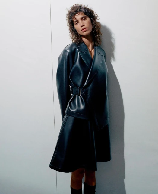 A woman in a black leather belted dress stands in a studio with a neutral expression. She is looking directly at the camera.