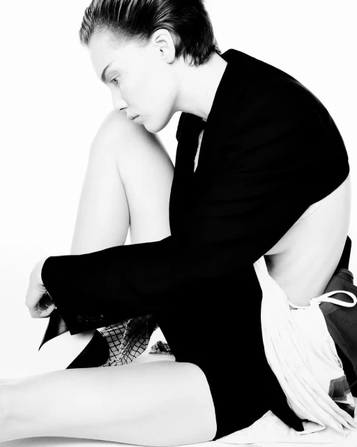 Portrait of a young adult woman in a black jacket sitting in a photo studio, with short hair and a neutral facial expression, against a white backdrop.