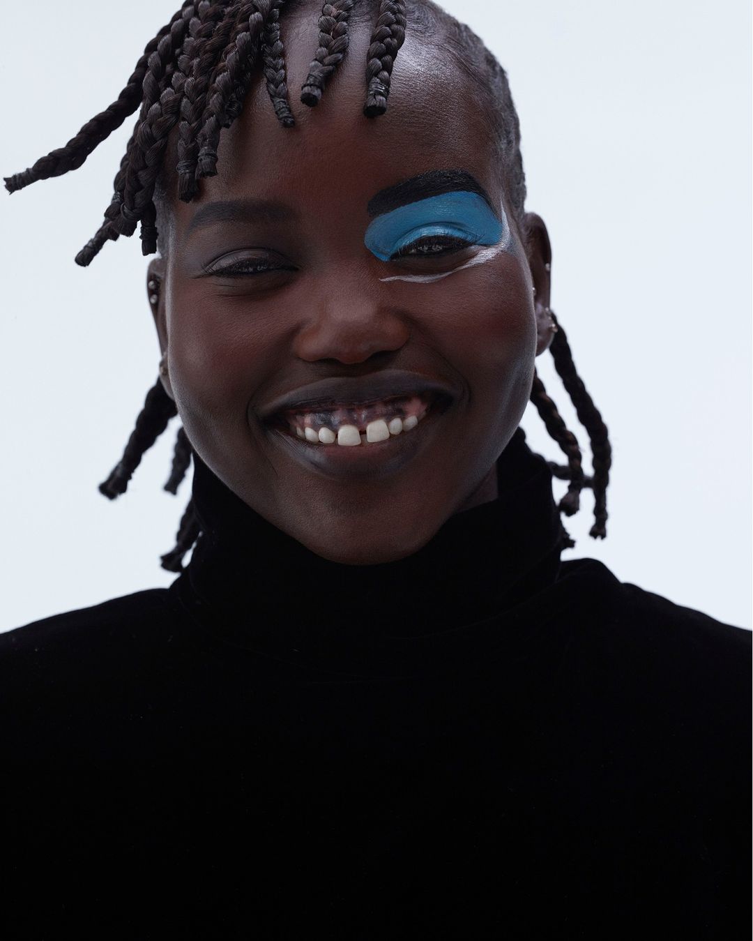 Close-up of a smiling young Black woman with dark complexion, braided hair, and bold blue eyeshadow on one eyelid against white.