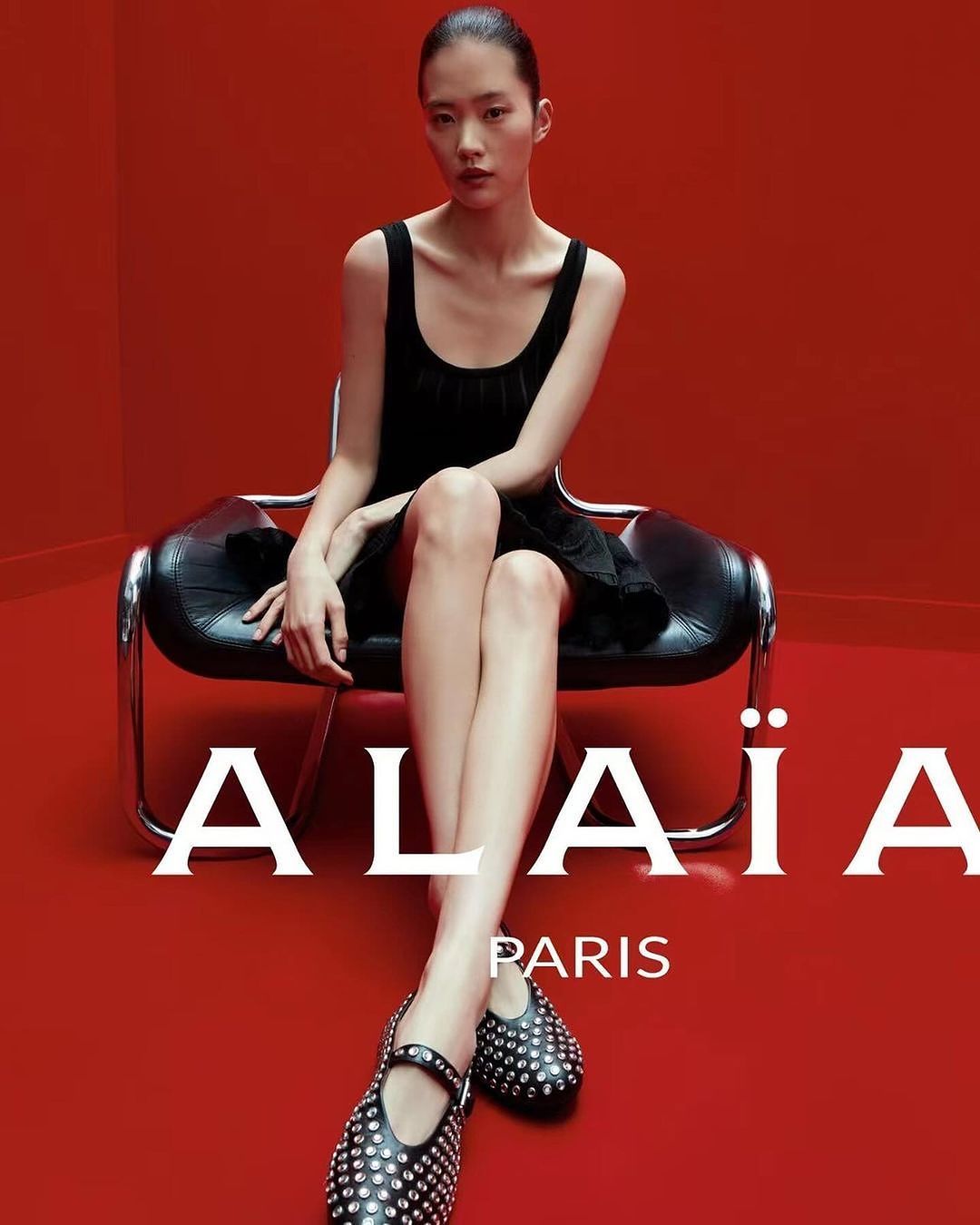 Fashion shoot of a slender Asian woman in a black dress and studded flats, seated on a chair against a bright red studio backdrop.