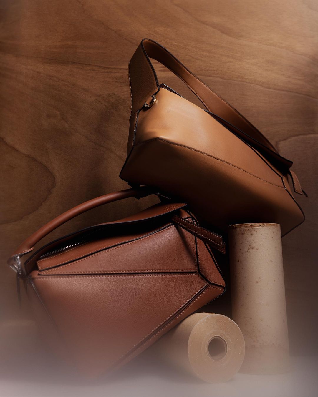 Two stacked brown leather handbags sit next to cylindrical props against a wood backdrop, creating a stylish, neutral product shot.