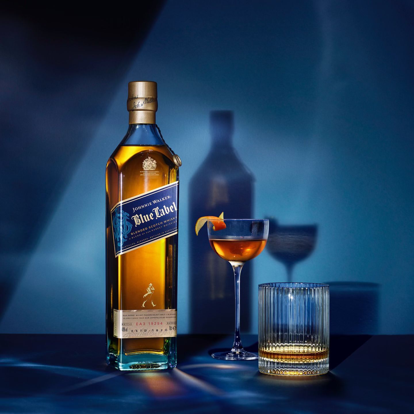 Johnnie Walker Blue Label bottle with glasses of amber liquid and orange peel garnish on a blue surface and blue background.