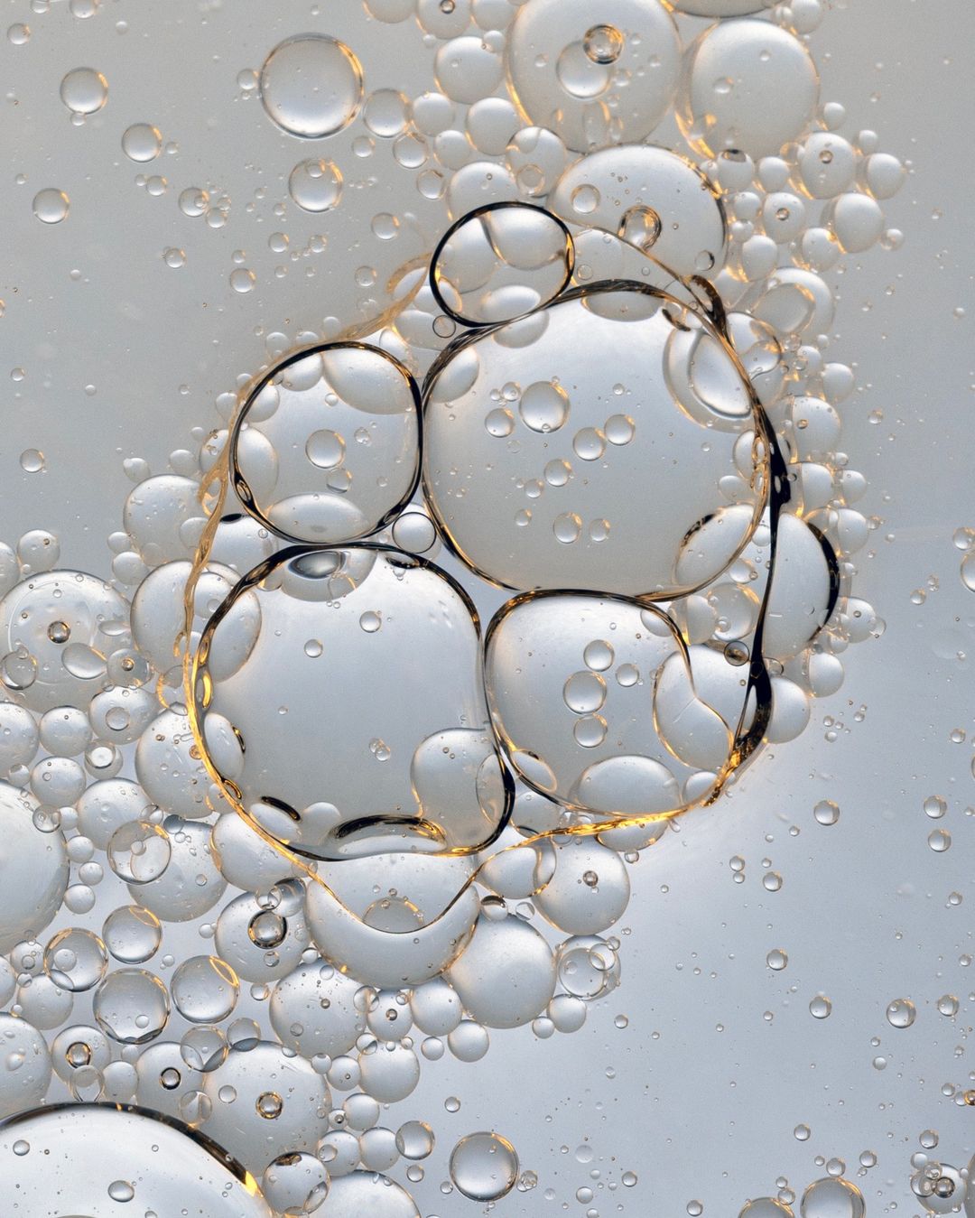 Close-up of liquid with translucent bubbles of varying sizes and shapes, some merging, against a pale background. Abstract photography.