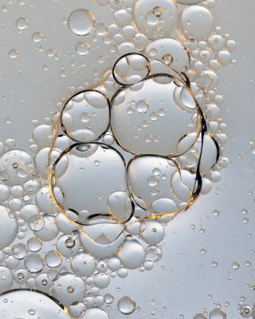 Close-up of liquid with translucent bubbles of varying sizes and shapes, some merging, against a pale background. Abstract photography.