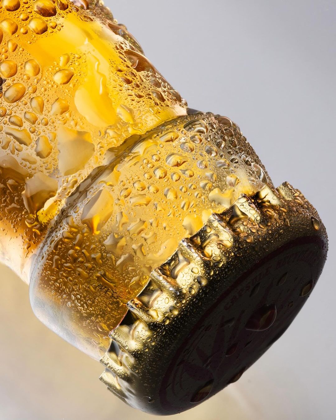 Close-up of a wet glass beer bottle neck with a metal cap, condensation droplets, and yellow liquid against a gradient background.