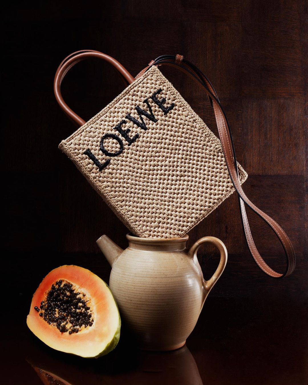 Woven straw Loewe bag with leather handle on a ceramic teapot next to half a papaya, against a dark brown textured background.
