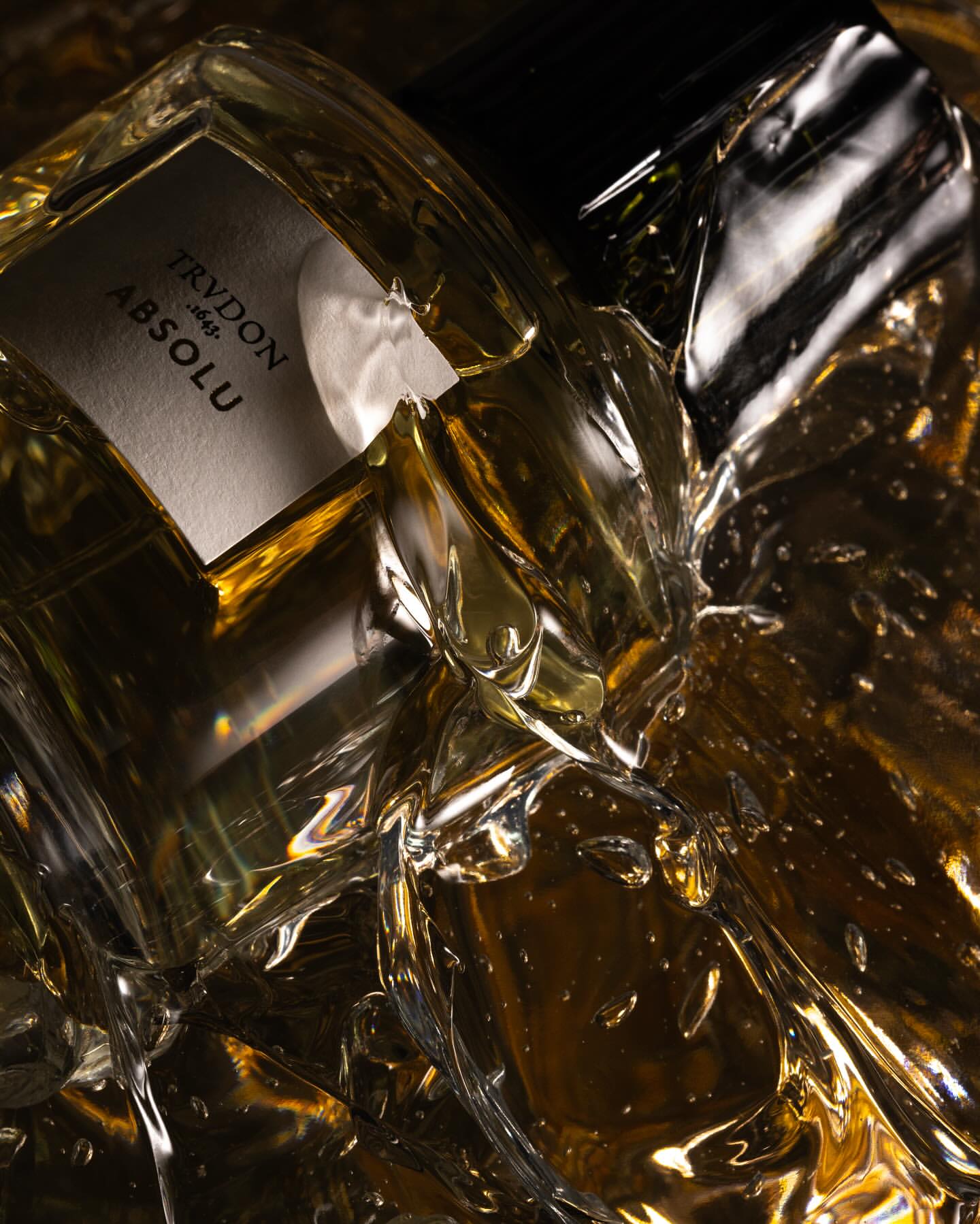 Close-up of a clear glass Trudon Absolu perfume bottle, surrounded by shiny, crinkled gold material in a dark studio setting.