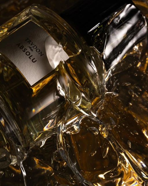 Close-up of a clear glass Trudon Absolu perfume bottle, surrounded by shiny, crinkled gold material in a dark studio setting.