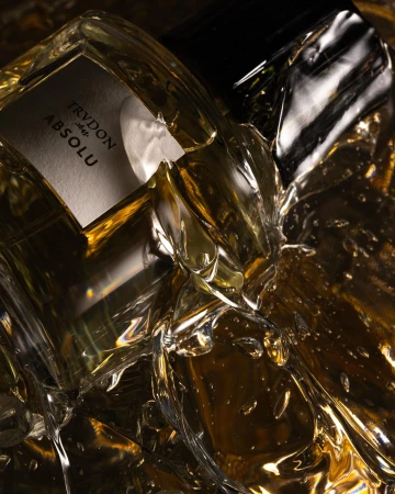 Close-up of a clear glass Trudon Absolu perfume bottle, surrounded by shiny, crinkled gold material in a dark studio setting.