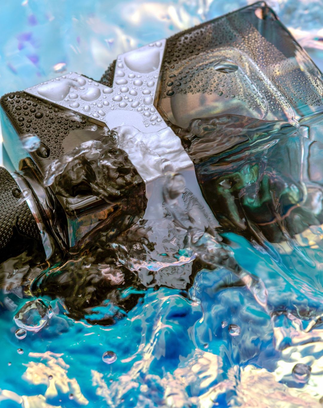 Close-up of an angular black perfume bottle with water droplets against a bright blue and white watery background.