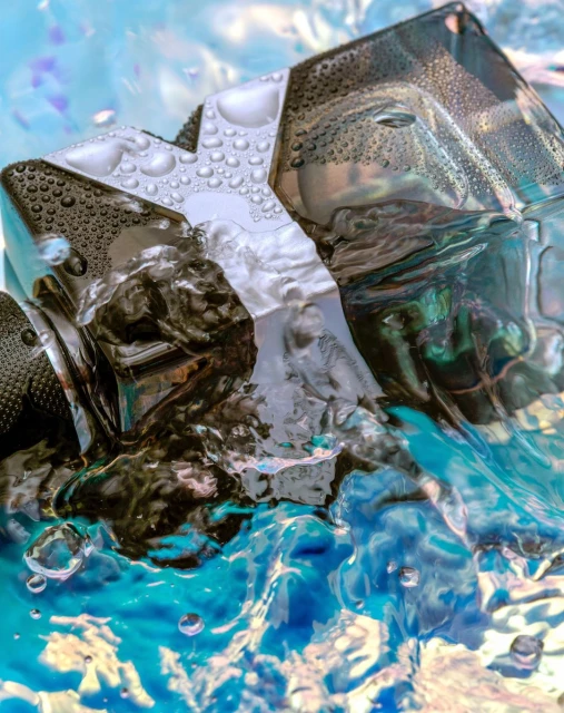 Close-up of an angular black perfume bottle with water droplets against a bright blue and white watery background.