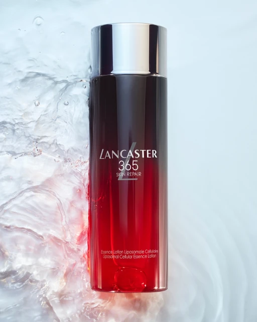 Lancaster 365 Skin Repair essence lotion bottle with black and red gradient, silver cap, and water splashes in a beauty shot.
