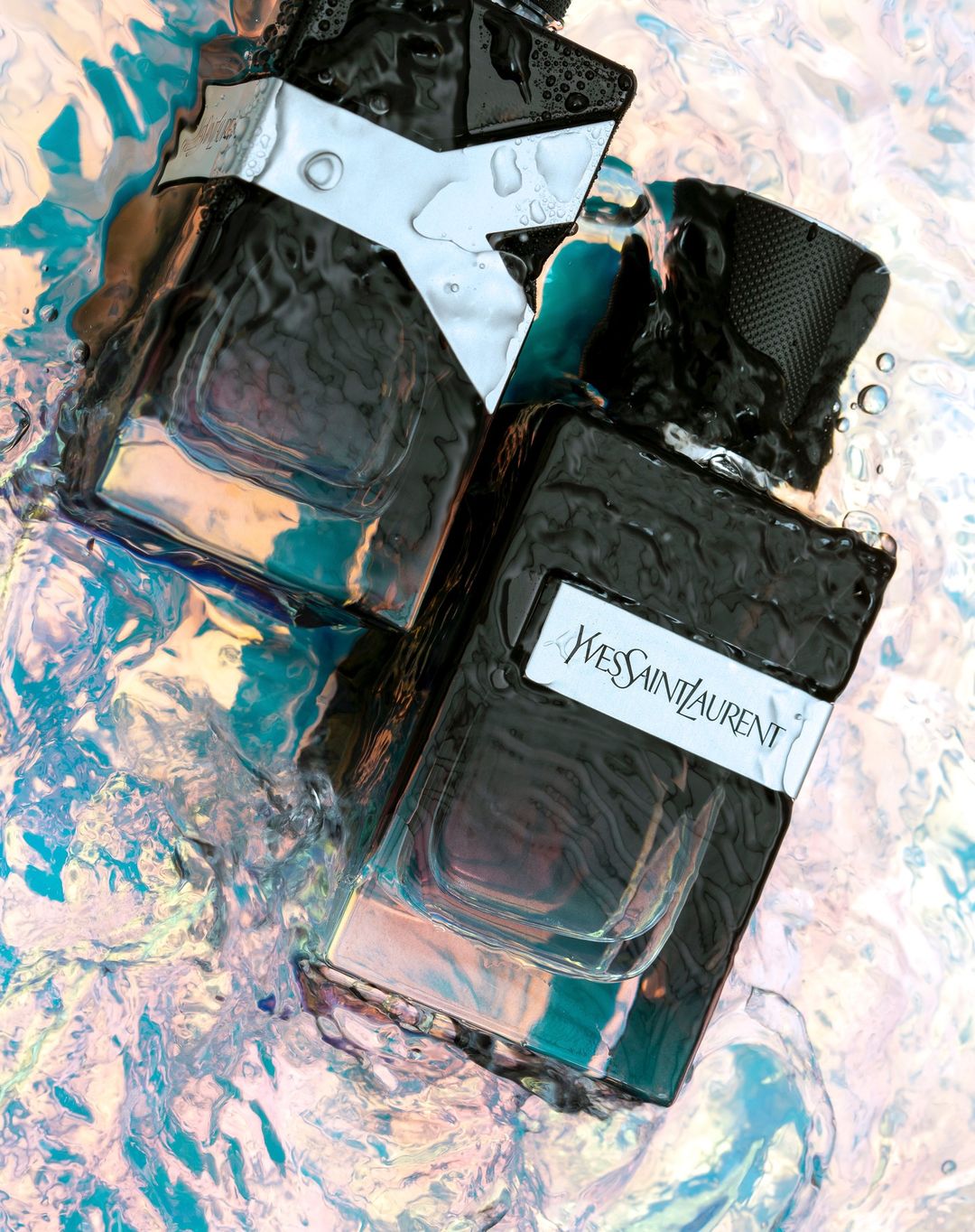 Two black bottles of Yves Saint Laurent cologne are submerged in water. The light reflects in shades of blue, pink, and white.