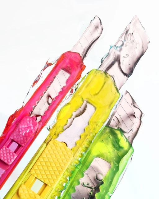 Eye-level studio shot of three vibrant box cutter knives in pink, yellow, and green, partially covered with clear liquid.