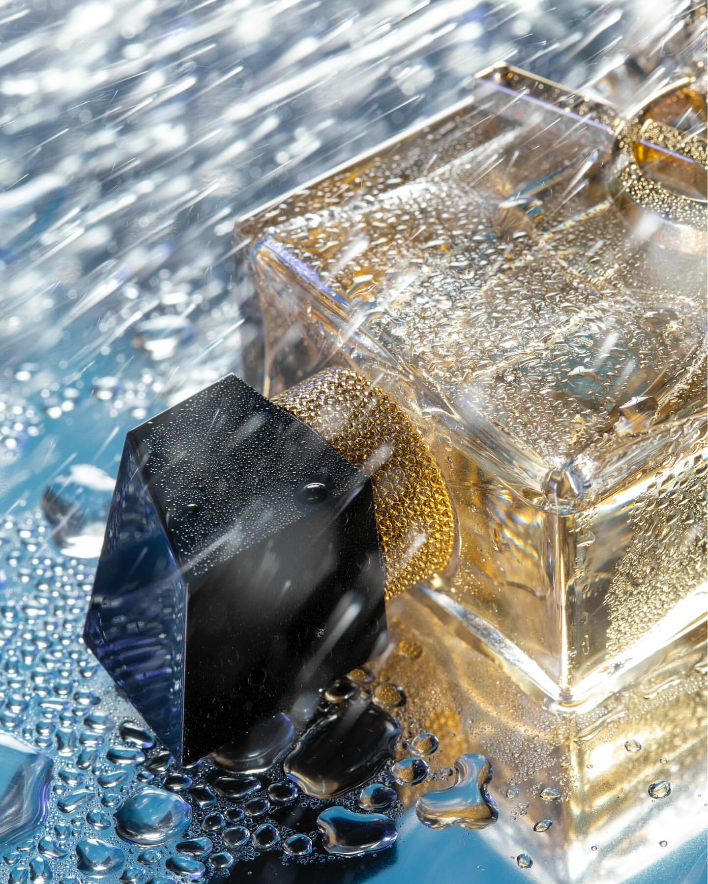 Close-up of a clear perfume bottle with a gold top and a black stopper, wet with droplets, against a blurred background.