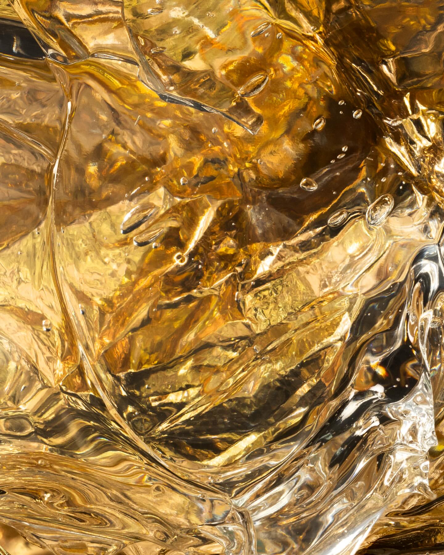 Close-up abstract photograph showcases textured amber liquid with rippling patterns, light reflections, and surface droplets.