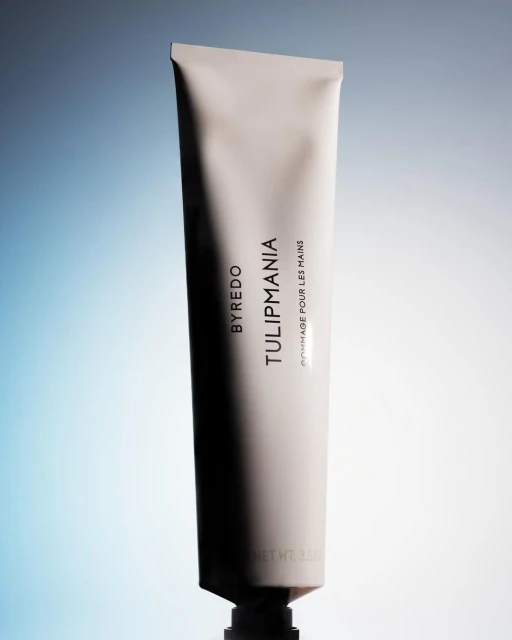 Byredo Tulipmania hand cream tube stands vertically, featuring gray and white gradient background, studio lighting, and reflective surface.