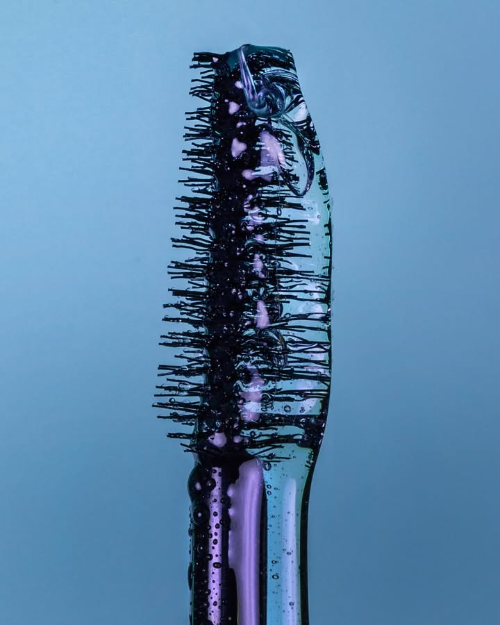 Macro shot of a mascara wand with visible clear serum on black bristles against a light blue background in a studio.