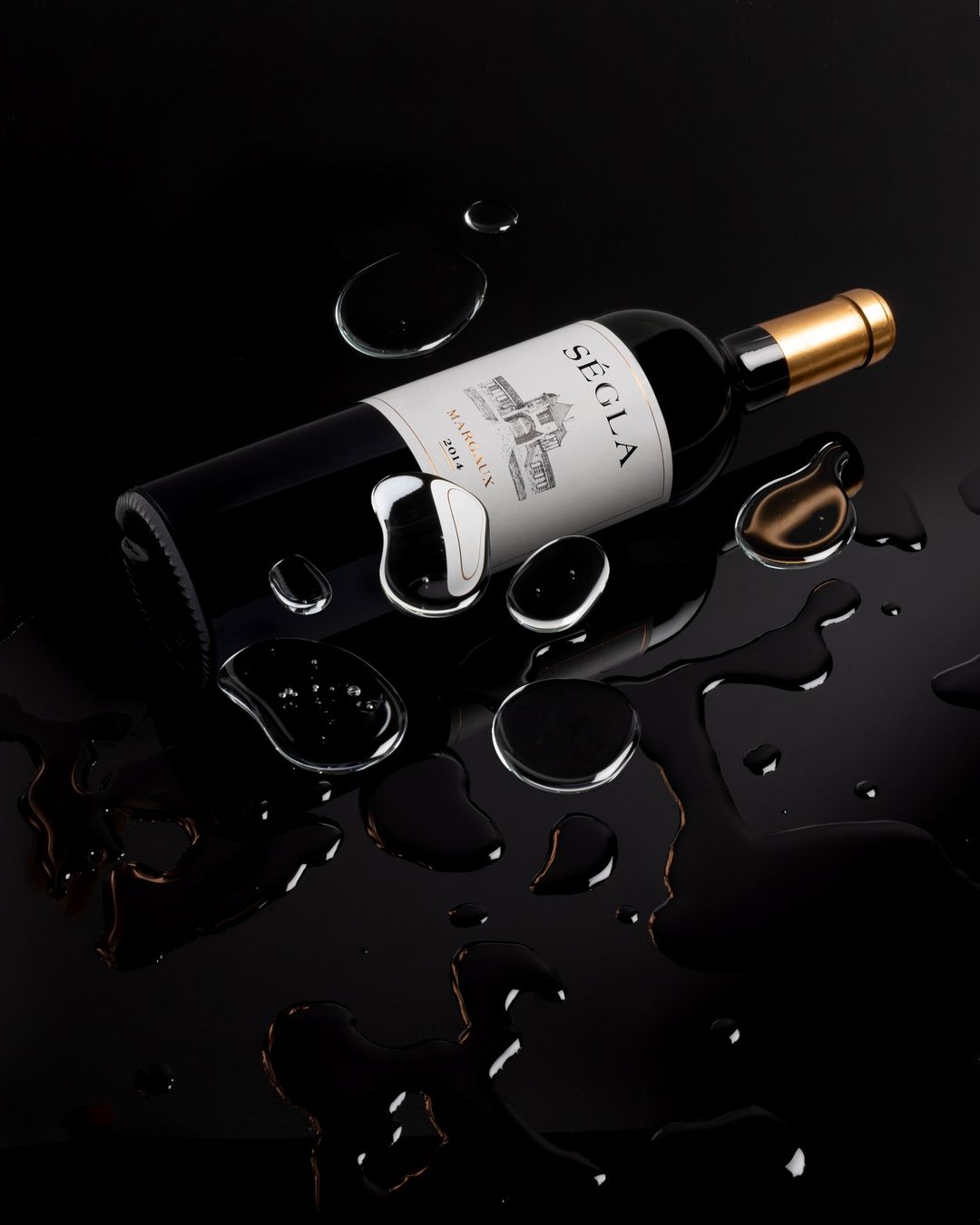 Studio shot of red wine bottle with gold cap, water droplets around it on shiny black surface creating reflections and dramatic effect.