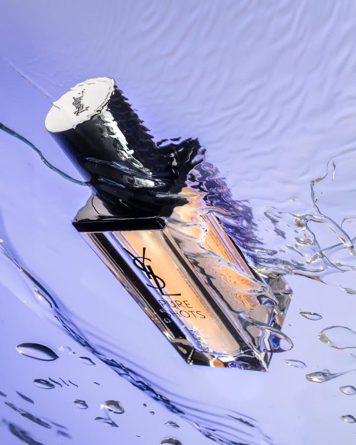 Close-up product shot of YSL Pure Shots perfume bottle submerged in water, highlighting its silver cap, clear glass, and amber liquid against a blue background.