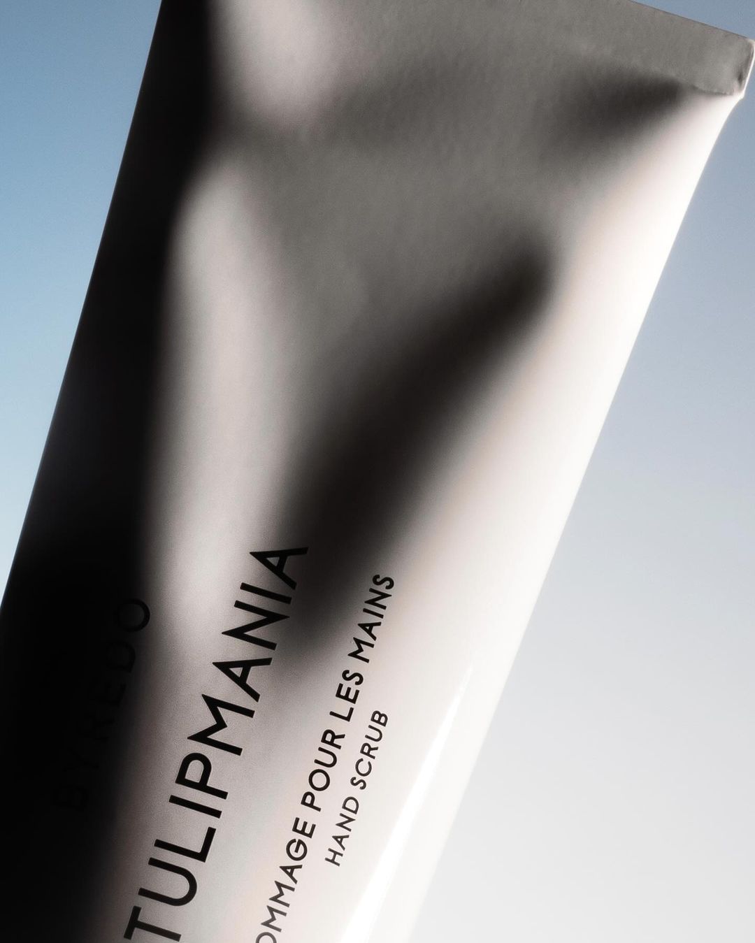 Close-up of a white tube of Tulipmania hand scrub against a soft blue and white gradient background, showcasing beauty product packaging.