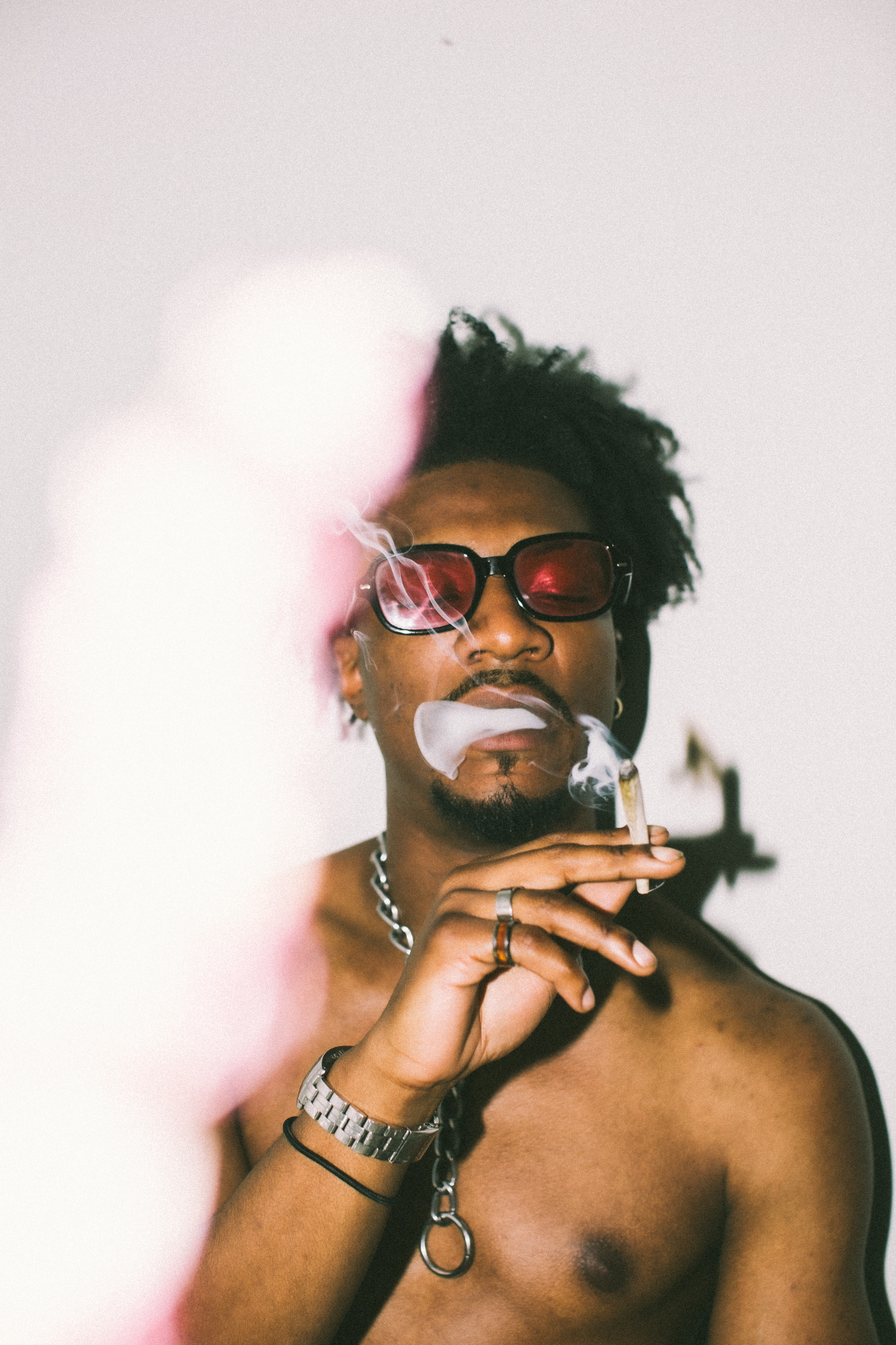 Portrait of a shirtless African American male smoking with smoke surrounding him. He wears red sunglasses, silver chain and watch. White background.