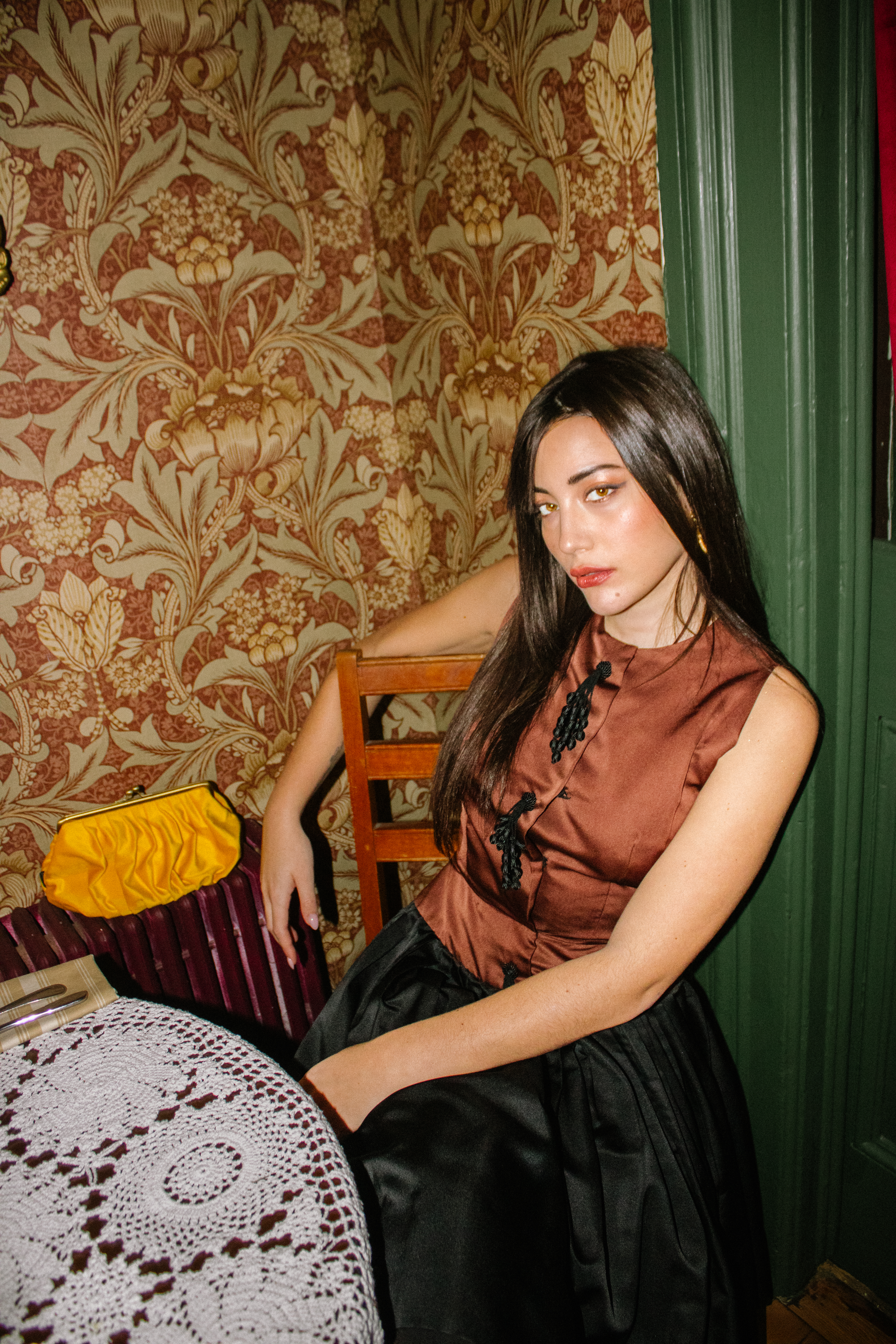 Elegant woman with long dark hair sits in vintage room with floral wallpaper wearing a brown top, black skirt, and golden earrings.