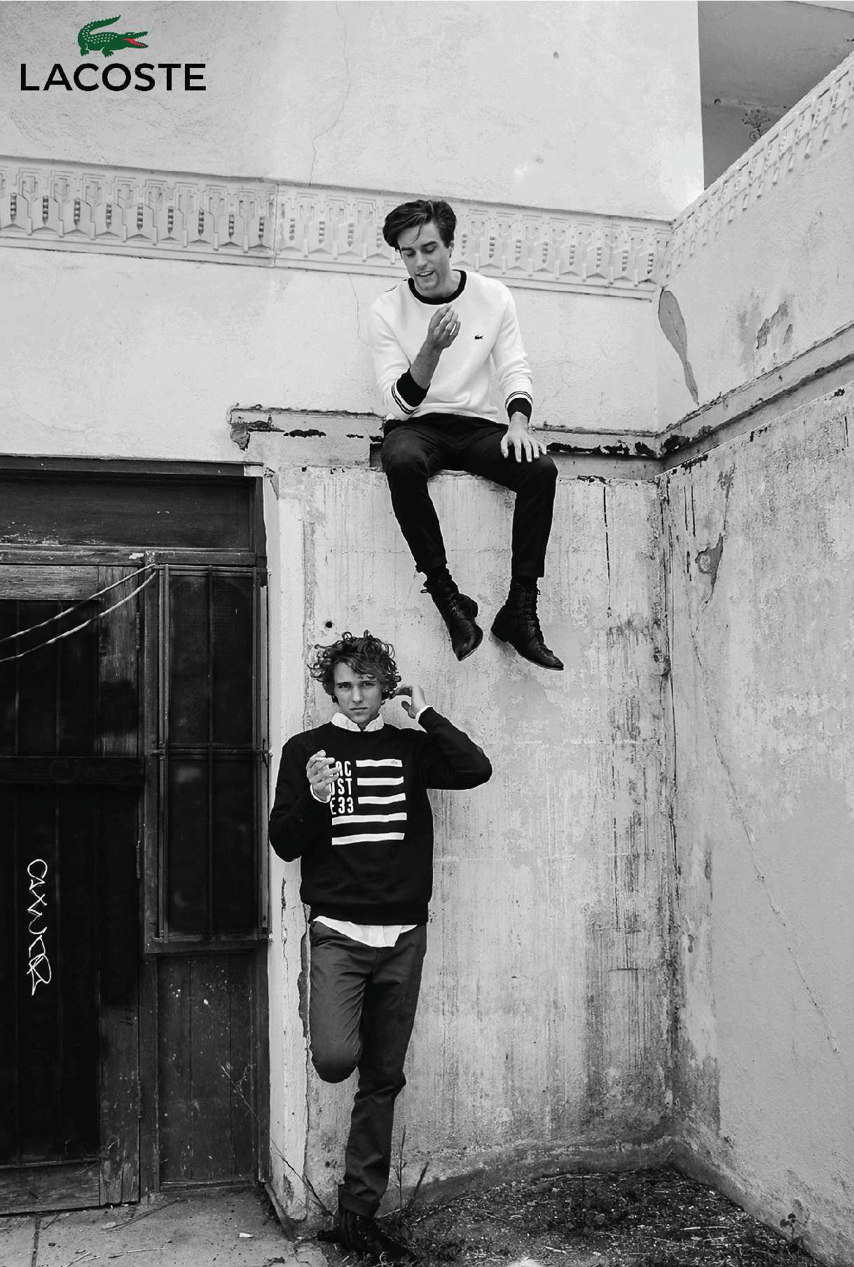 Black and white fashion shot of two young men in casual outfits posing against an old wall, featuring the Lacoste brand logo.