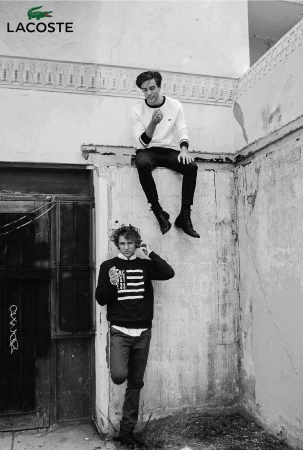Black and white fashion shot of two young men in casual outfits posing against an old wall, featuring the Lacoste brand logo.
