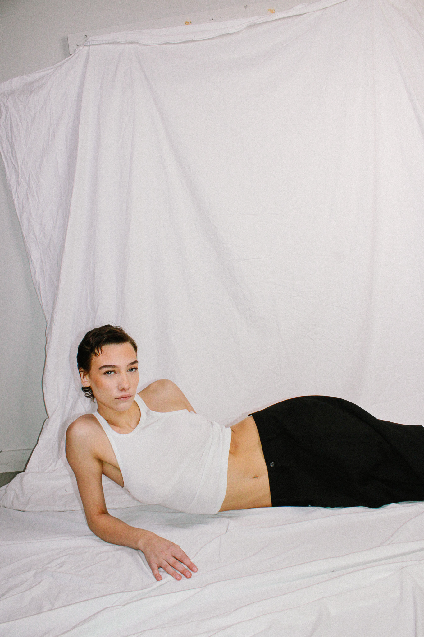 Adult female lying on a white fabric backdrop in a white top and dark pants. Simple studio style with natural-look skin.