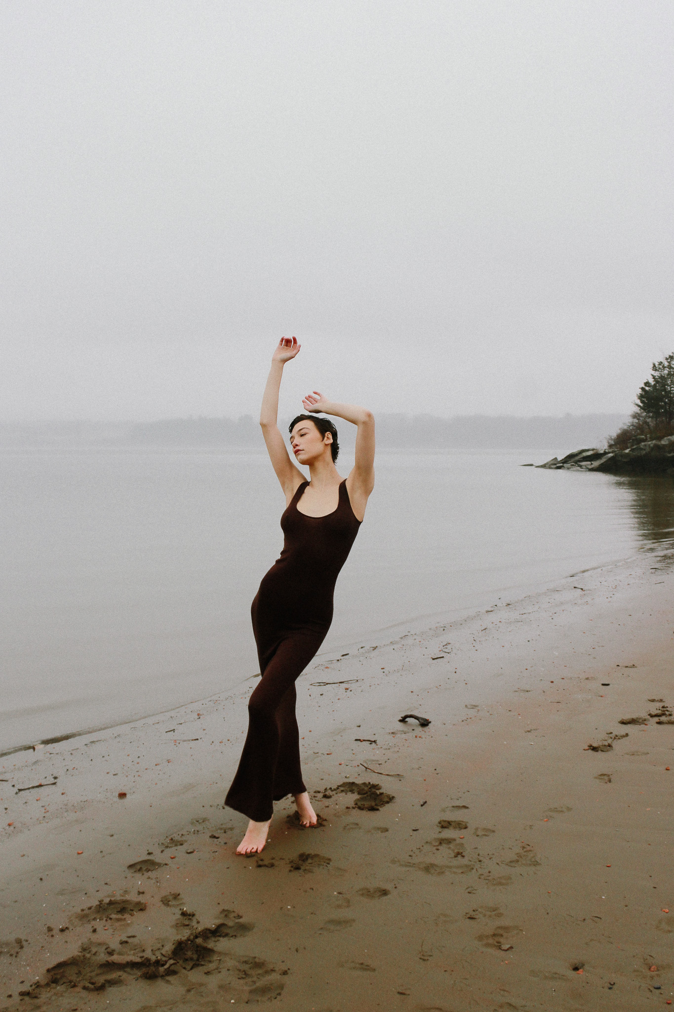 Young woman in brown jumpsuit poses on a sandy beach in front of water under a cloudy sky, striking a graceful pose with arms raised.