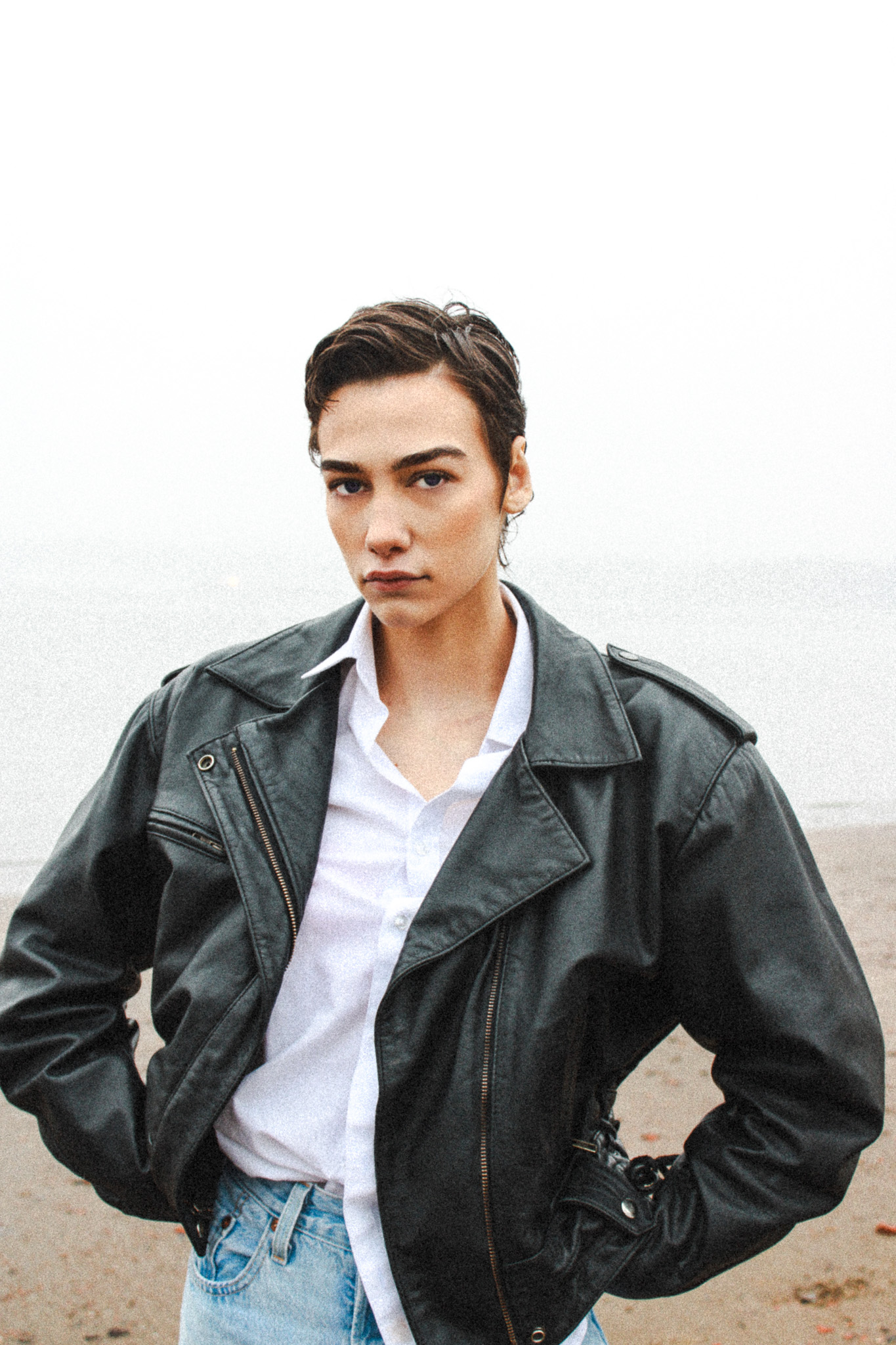 Adult woman with short hair in a white shirt and black leather jacket poses outdoors against a blurred background on a cloudy day.