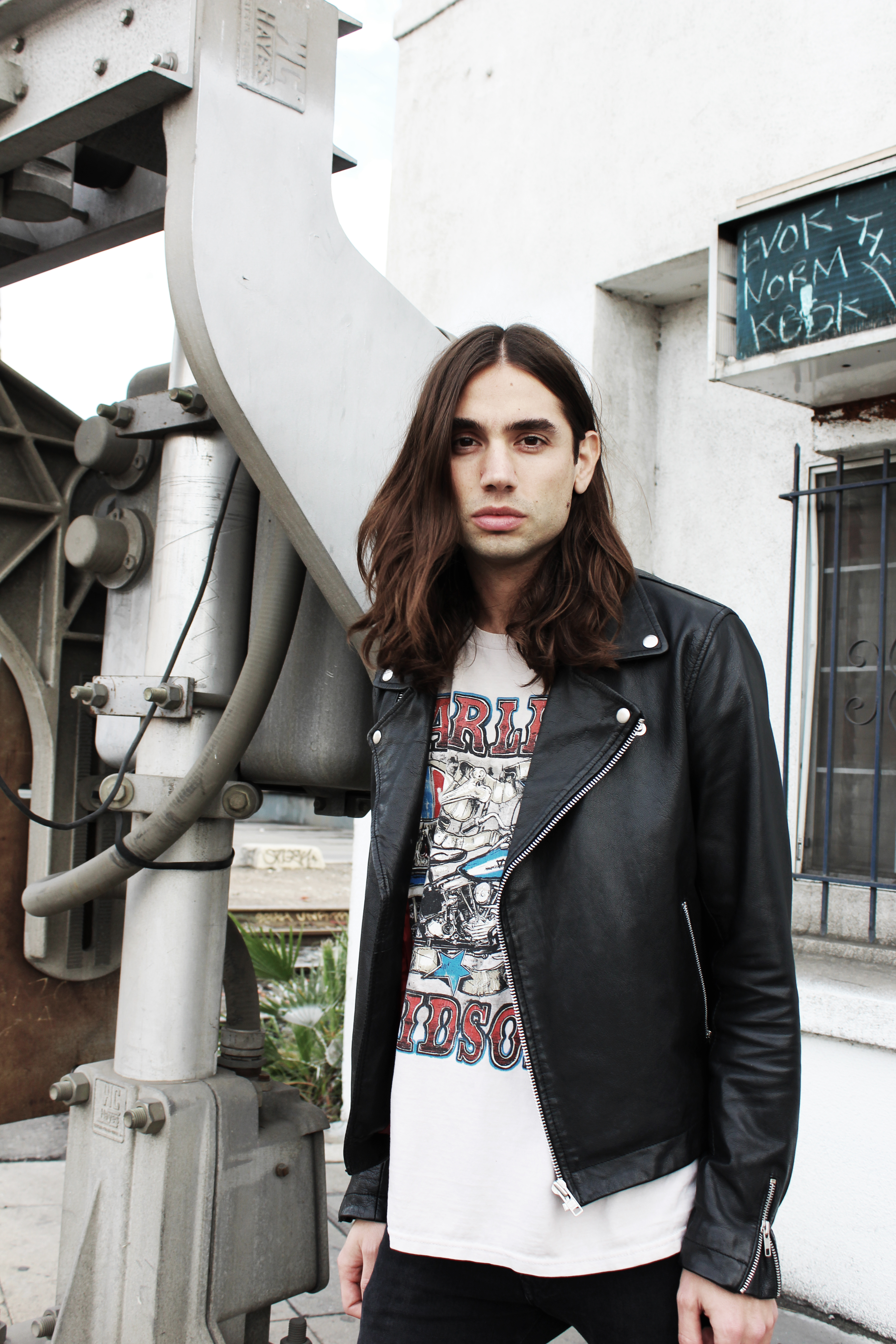 A slender adult wearing a black leather jacket and T-shirt poses outdoors against an industrial backdrop, captured in a medium shot.