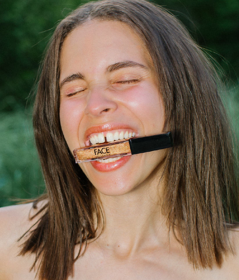 Woman with brown hair poses with closed eyes and a slight smile, holding a tube of gold lip gloss in her teeth outdoors.