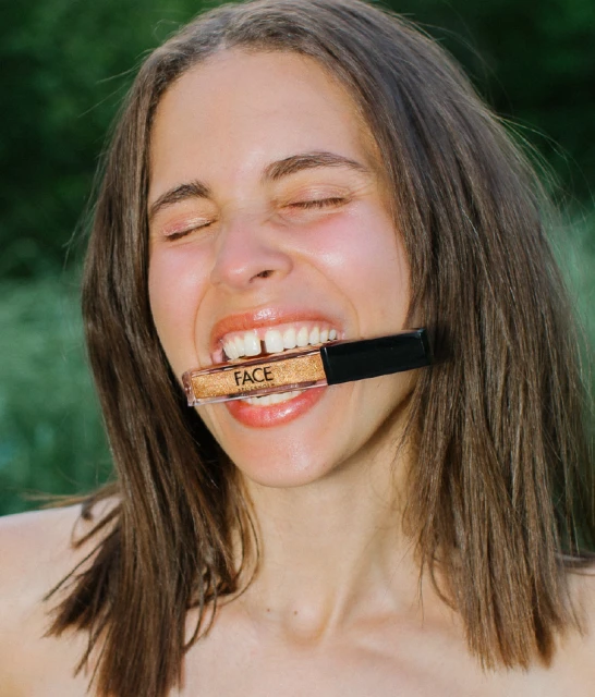 Woman with brown hair poses with closed eyes and a slight smile, holding a tube of gold lip gloss in her teeth outdoors.