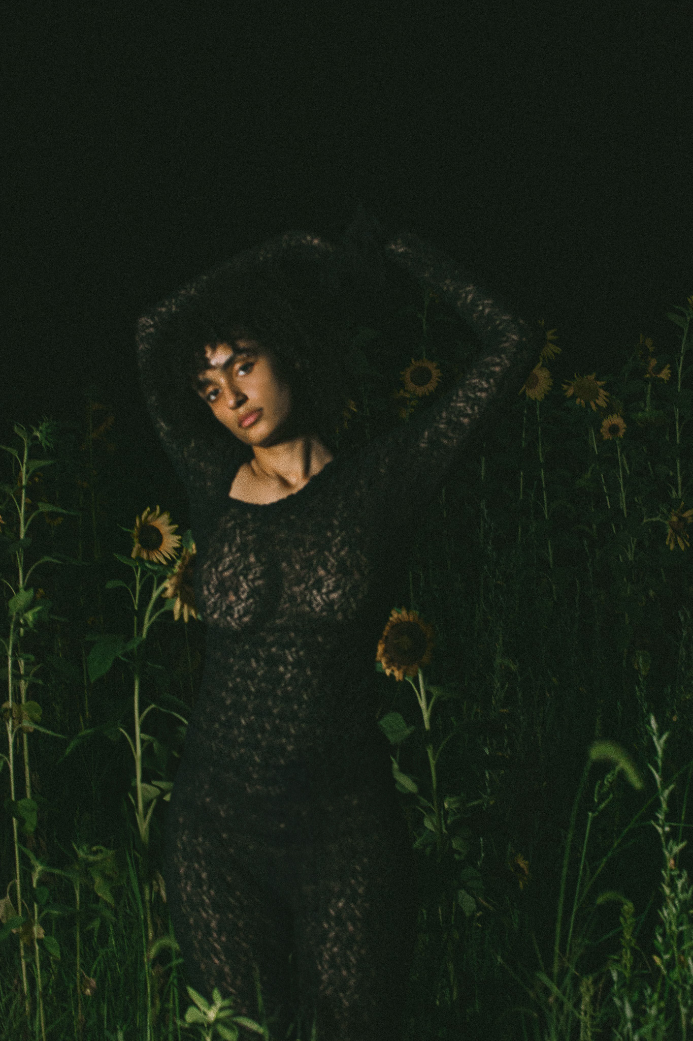 Woman in a floral dress stands in a field of sunflowers, illuminated in dark, moody lighting, creating a striking natural fashion portrait.