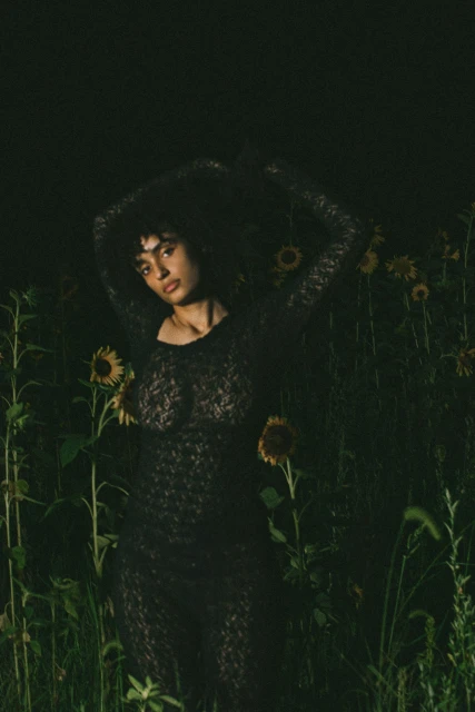 Woman in a floral dress stands in a field of sunflowers, illuminated in dark, moody lighting, creating a striking natural fashion portrait.
