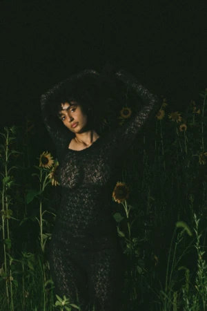 Woman in a floral dress stands in a field of sunflowers, illuminated in dark, moody lighting, creating a striking natural fashion portrait.
