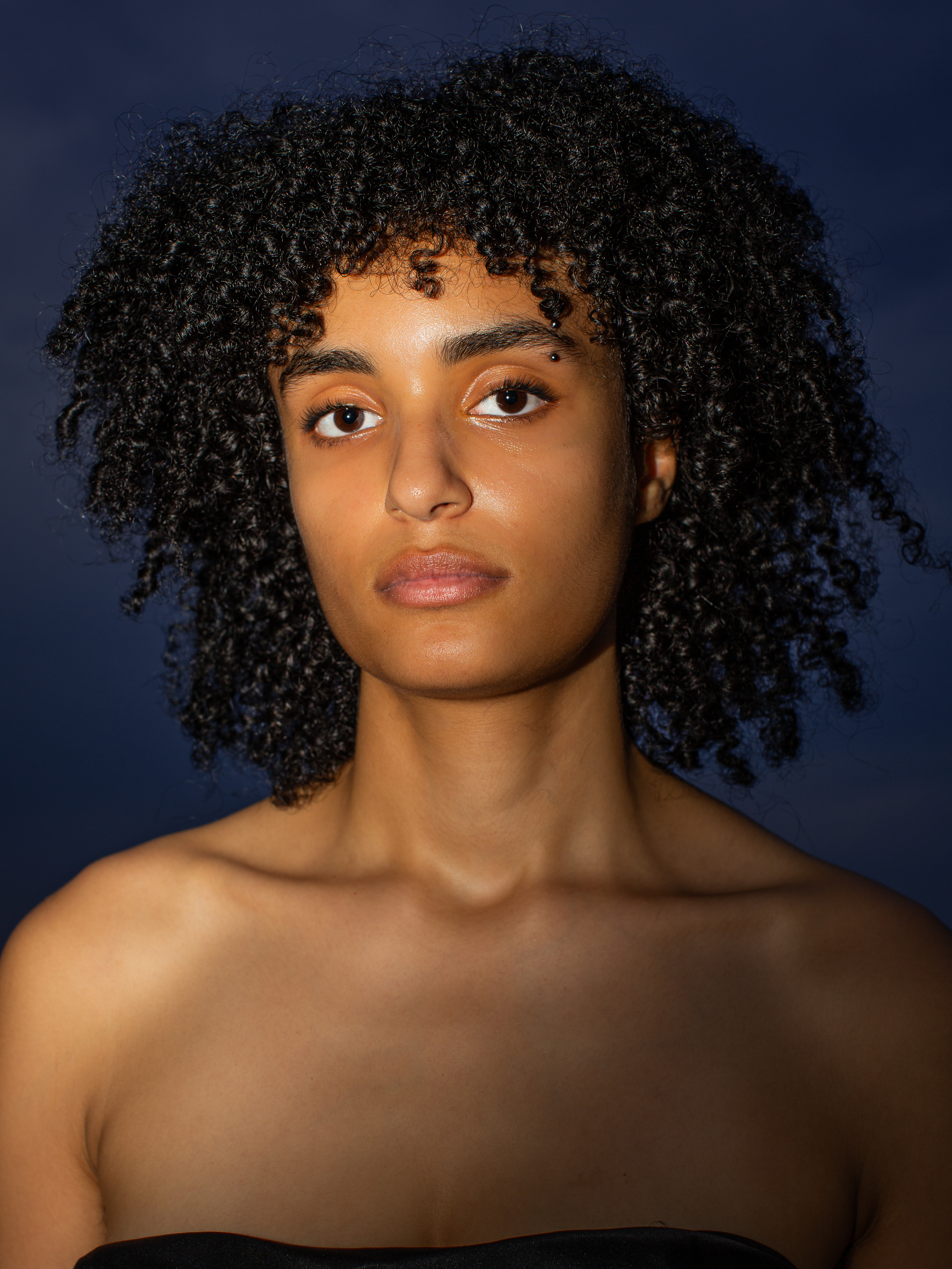 Portrait of a young, elegant woman with dark curly hair and minimal makeup against a dark, moody blue background.