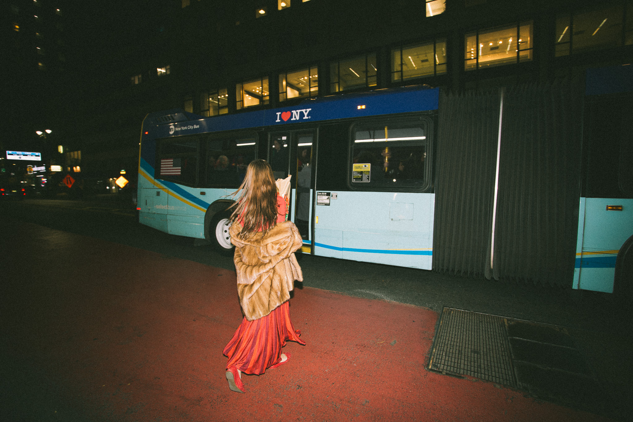 Woman in a red dress and fur coat walks towards a blue New York City bus at night, with warm-toned light from buildings in the background.