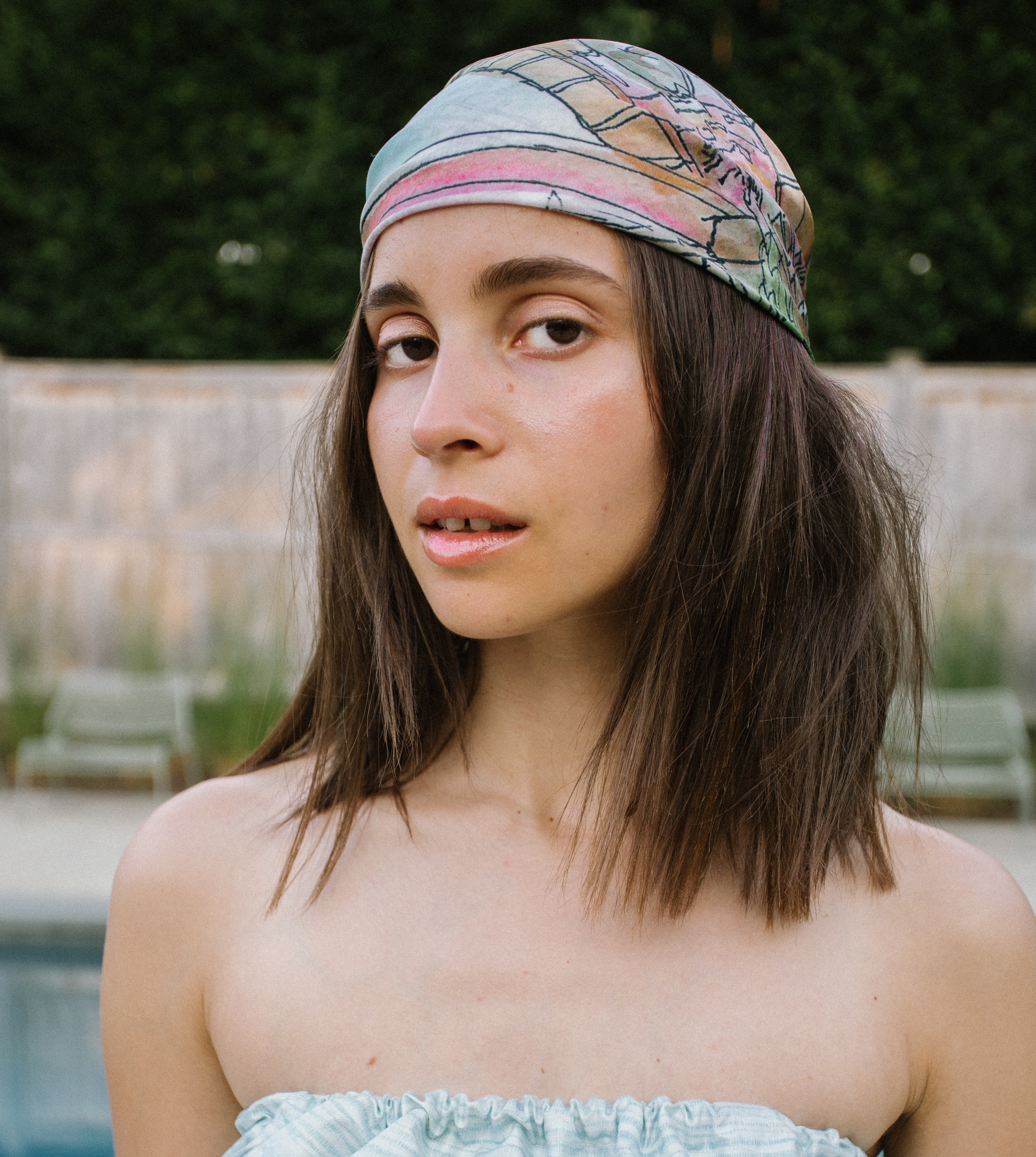 Portrait of a young woman with brown hair wearing a patterned headscarf, posing outside by a pool and green hedge in natural light.