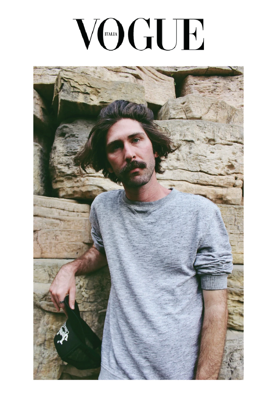 Man with a mustache wearing a grey crewneck sweatshirt leaning against a textured stone wall while holding a black cap.
