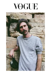 Man with a mustache wearing a grey crewneck sweatshirt leaning against a textured stone wall while holding a black cap.