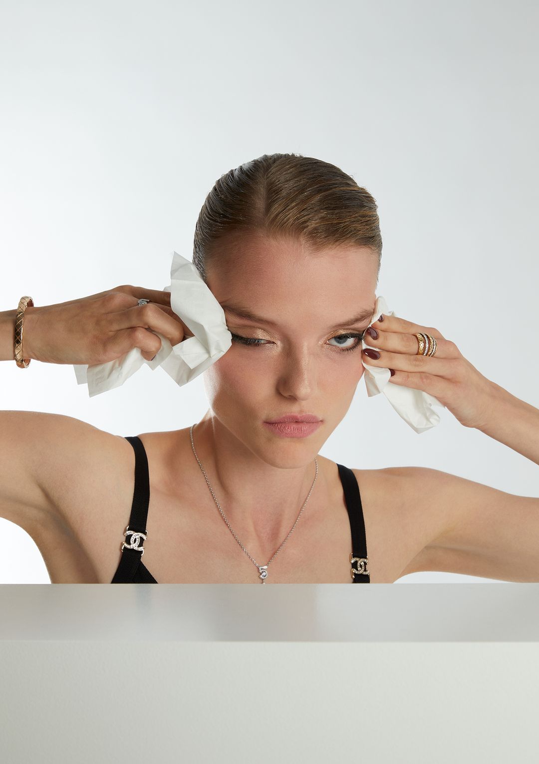 Portrait of a young adult woman in a black top dabbing face with white tissues against a white background, accessorized with jewelry.