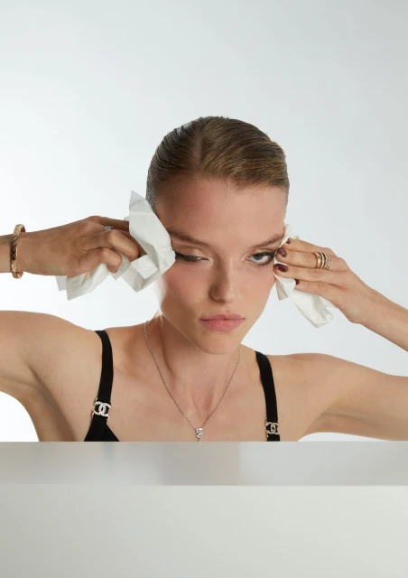 Portrait of a young adult woman in a black top dabbing face with white tissues against a white background, accessorized with jewelry.